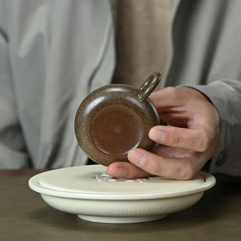 A hand holding a small brown ceramic cup with a loop handle above a white ceramic lid or saucer, with a person in a gray jacket blurred in the background.

