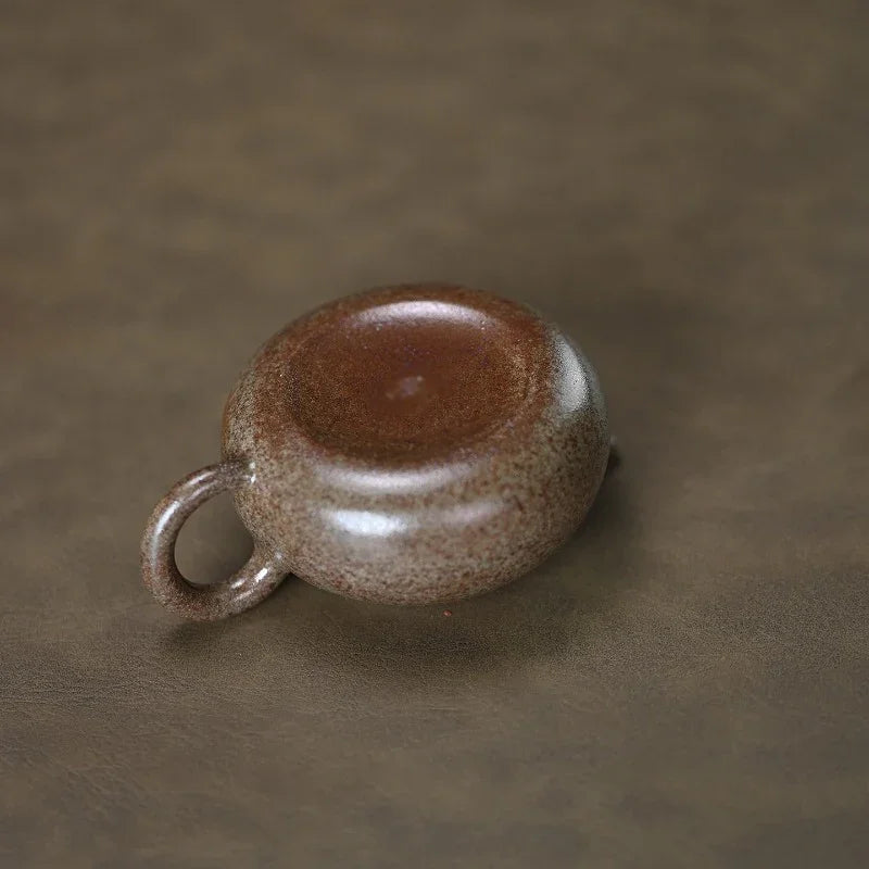 A small ceramic cup with a rounded body and a loop handle, shown from a top-down angle on a textured brown surface. The cup has a speckled brown and beige glaze with a glossy center.

