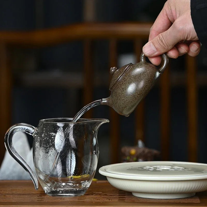 A hand holds a small speckled dark brown teapot pouring water into a clear glass pitcher, with a white ceramic lid resting on a wooden table in a tea setting.

