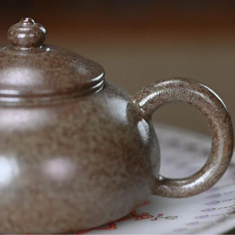 Close-up of a speckled ceramic teapot with a rounded handle and lid, shown on a patterned surface with a blurred warm background.

