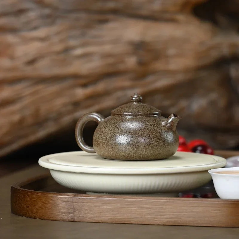 A speckled brown ceramic teapot with a lid sits on a round white saucer, placed on a wooden tray with a blurred rustic wood background.

