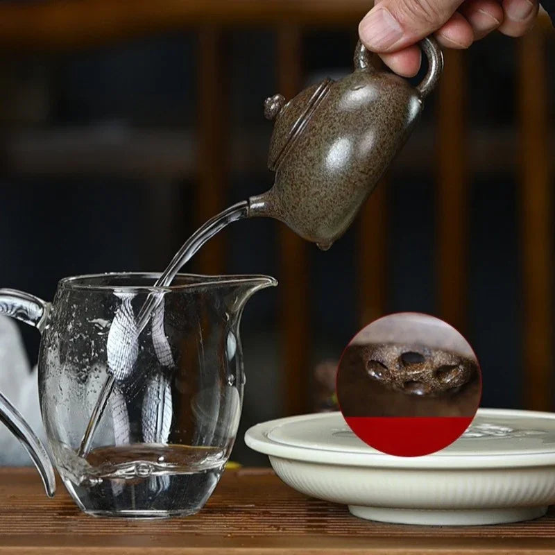 A hand pours water from a speckled brown ceramic teapot into a clear glass pitcher. A circular inset shows a close-up of the teapot's handle and spout.

