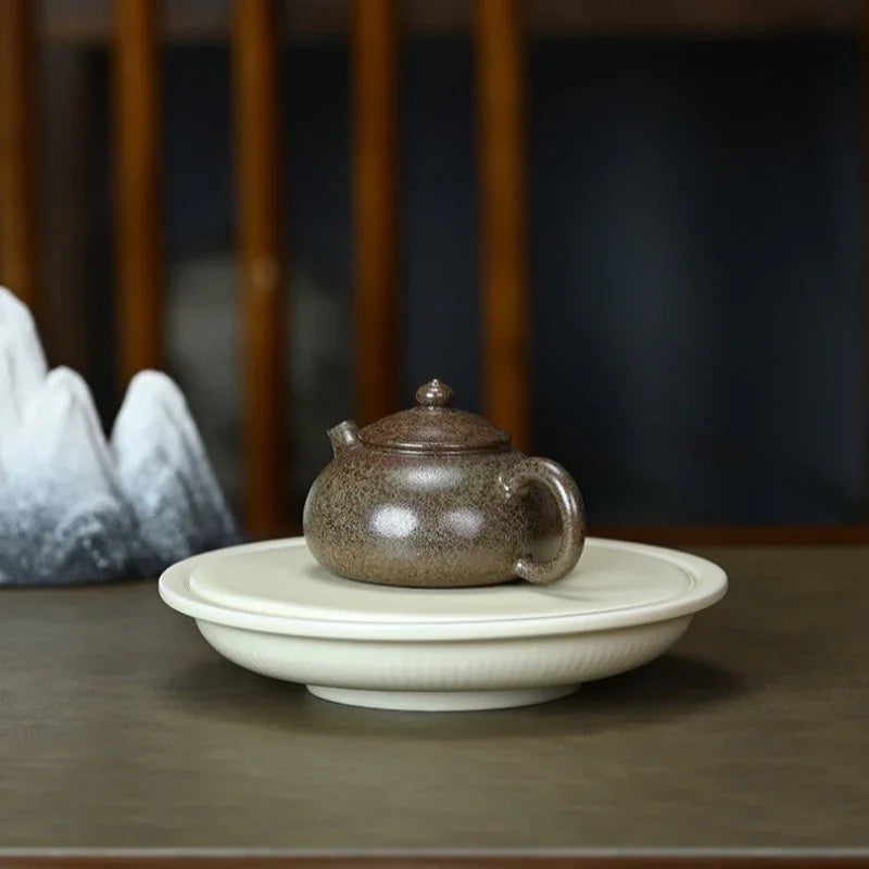A small speckled brown ceramic teapot sits centered on a round white saucer or tray, placed on a dark wooden table with a softly blurred indoor background.

