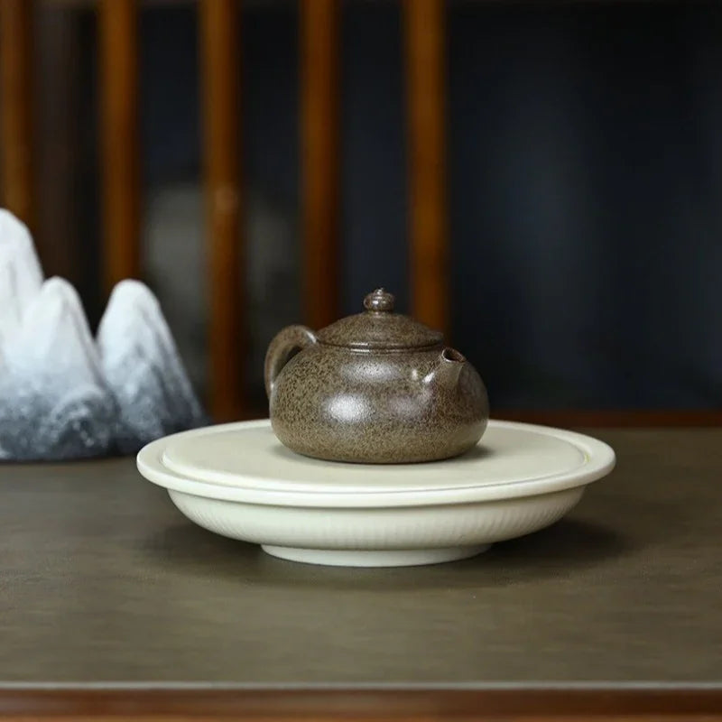 A small speckled brown ceramic teapot sits centered on a round white saucer, placed on a dark wooden table with a softly blurred indoor background.

