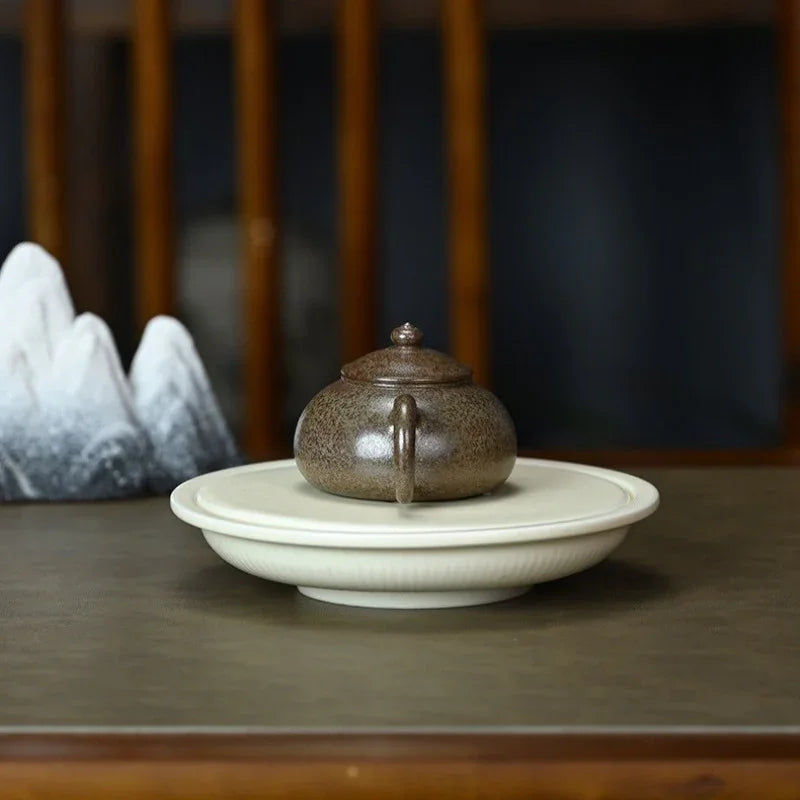 A small speckled brown ceramic teapot sits centered on a round white saucer, placed on a dark wooden table with a softly blurred indoor background.

