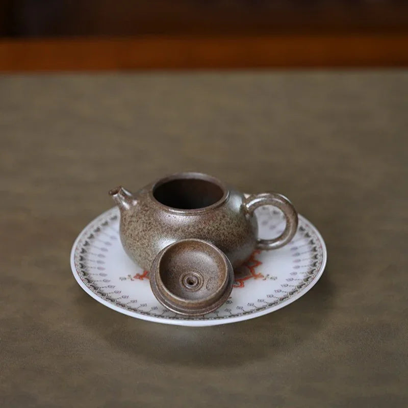 A small speckled ceramic teapot with its lid off, resting on a white saucer with a delicate red and gold pattern, set against a neutral brown background.

