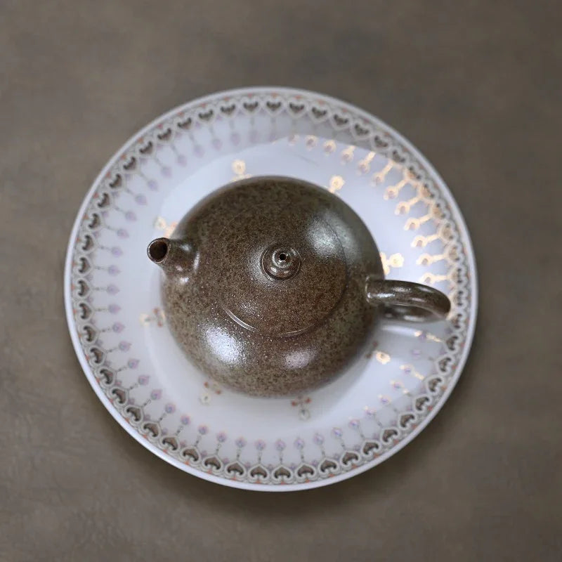 A dark brown speckled ceramic teapot resting on a white saucer with a delicate scalloped border, set against a neutral gray background.

