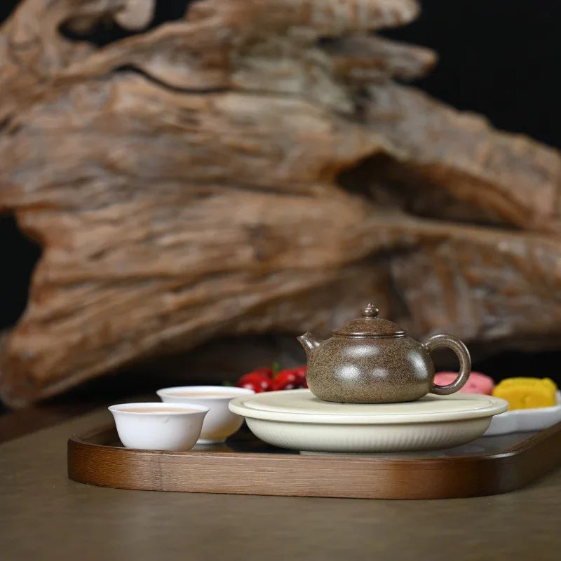 A ceramic tea set arranged on a wooden tray, featuring a speckled brown teapot, two small white cups, stacked cream-colored plates, and a blurred decorative wood background.

