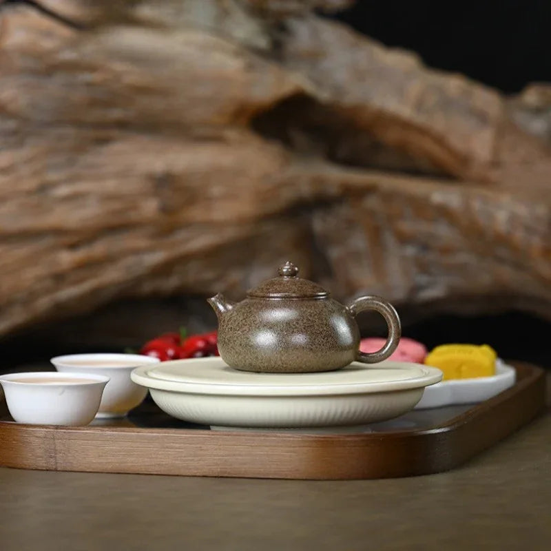 A small speckled brown ceramic teapot sits on a round cream-colored saucer, arranged on a wooden tray with white teacups and a blurred decorative wood background.

