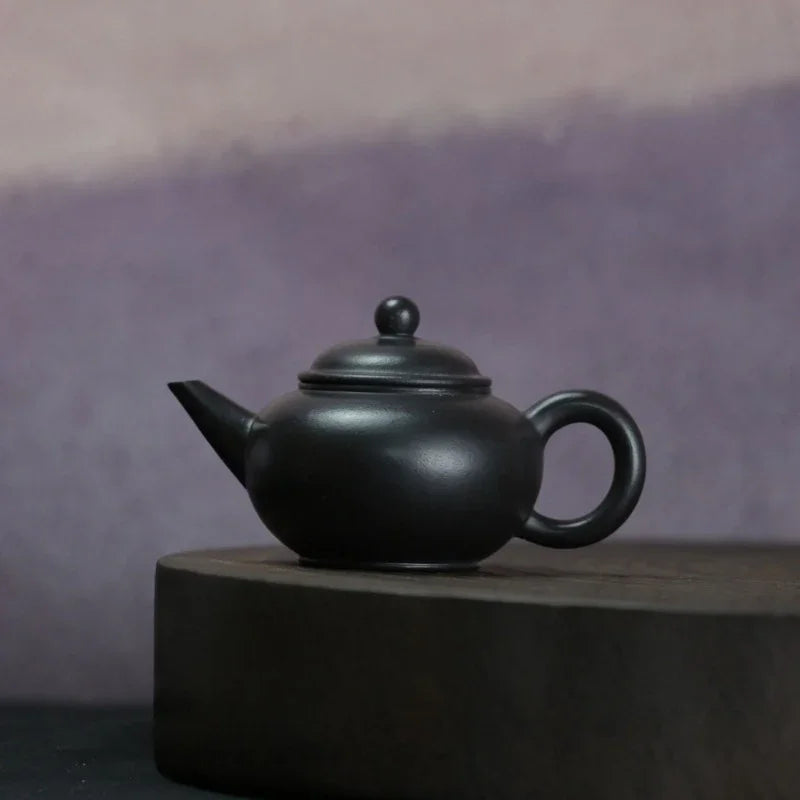 A matte black ceramic teapot with a rounded body, short spout, and loop handle, displayed on a dark round pedestal against a soft purple background.

