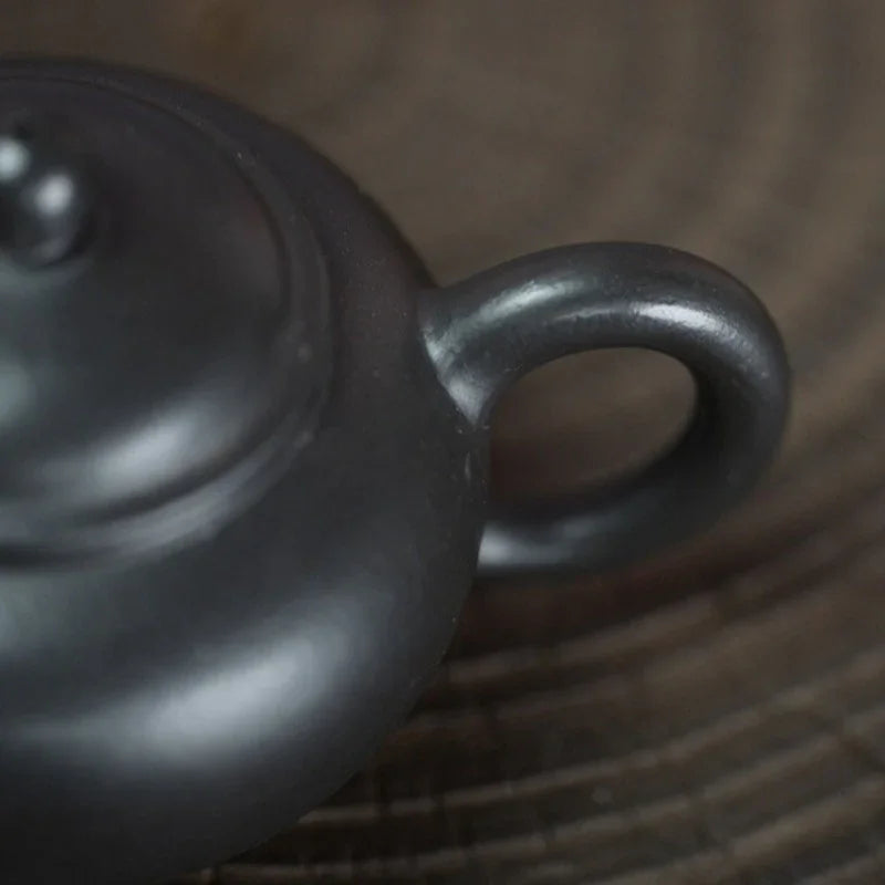 Close-up of a dark gray ceramic teapot with a loop handle, resting on a textured brown wooden surface.

