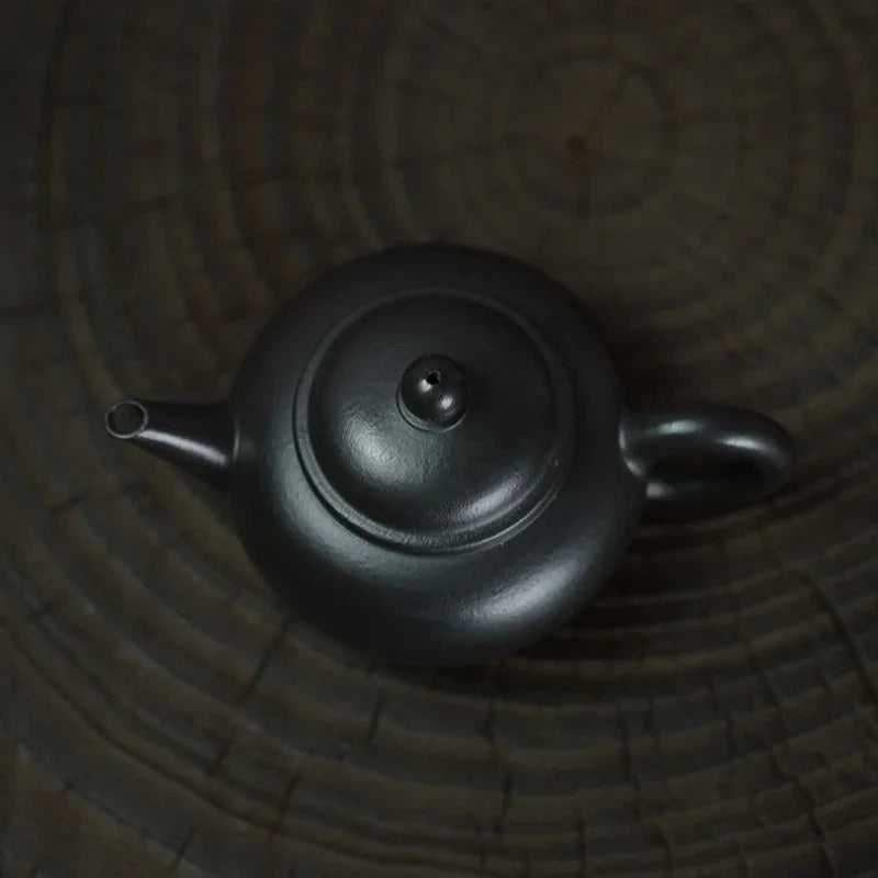 A dark charcoal black cast iron teapot with a rounded body, lid, and loop handle, resting on a dark textured wooden surface.

