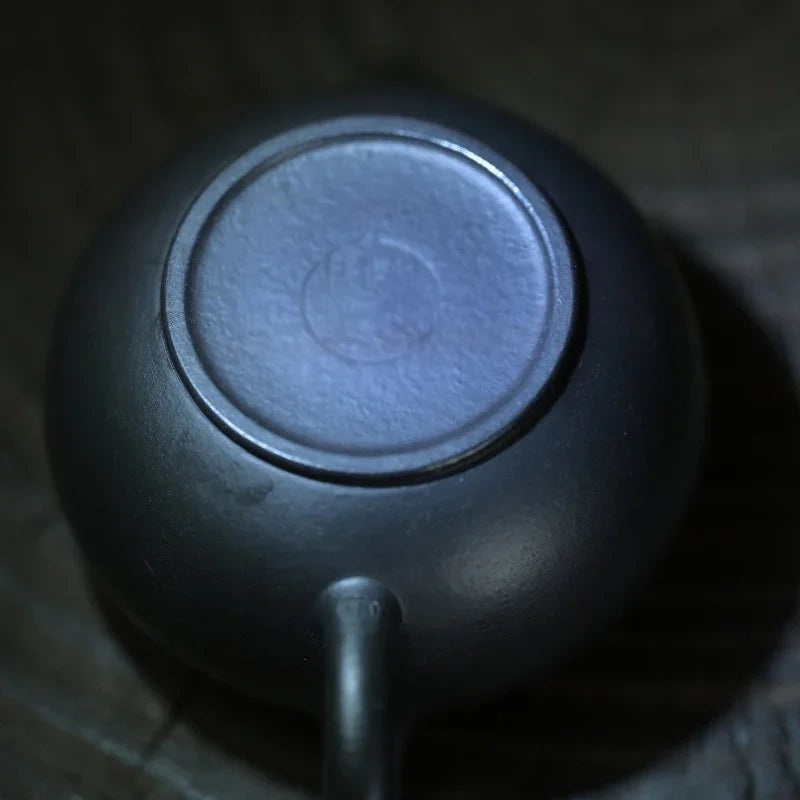 Close-up of the bottom of a dark ceramic teapot, showing the circular base and a small handle protruding downward, photographed against a dark wooden surface.

