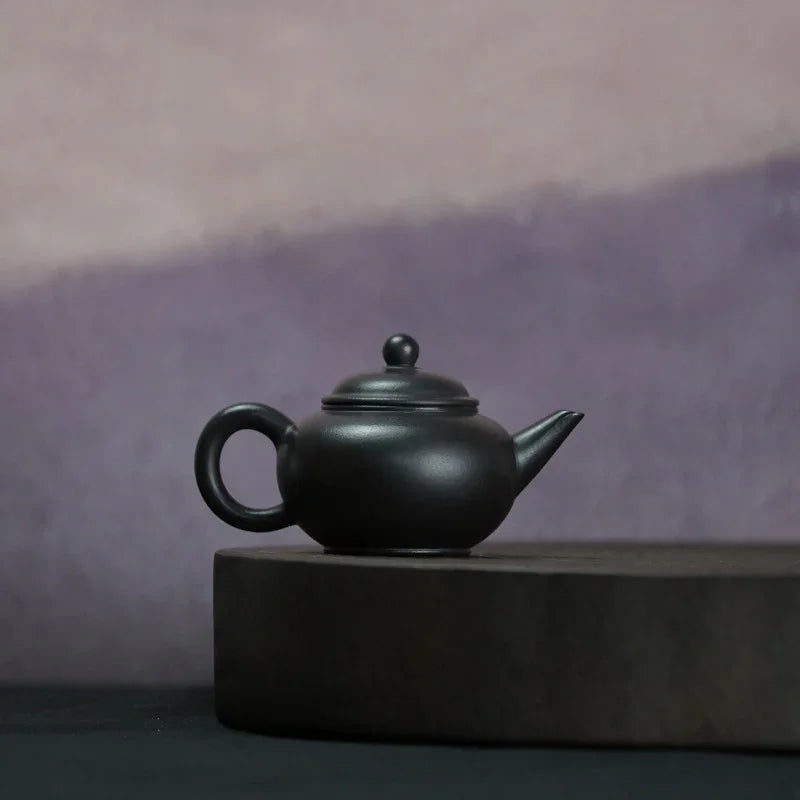 A matte black ceramic teapot with a rounded body, loop handle, and spout, displayed on a dark rectangular pedestal against a soft purple and mauve gradient background.

