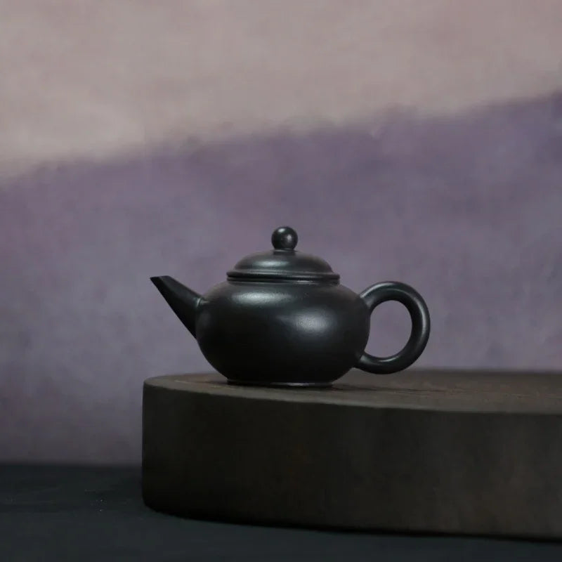 A matte black ceramic teapot with a rounded body, loop handle, and small spout, displayed on a round dark pedestal against a soft purple-gray background.

