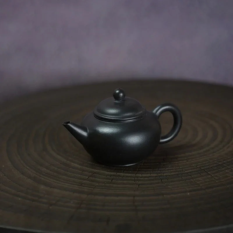 A small matte black ceramic teapot with a rounded body, curved spout, and loop handle, placed on a dark round wooden tray against a soft purple-gray background.

