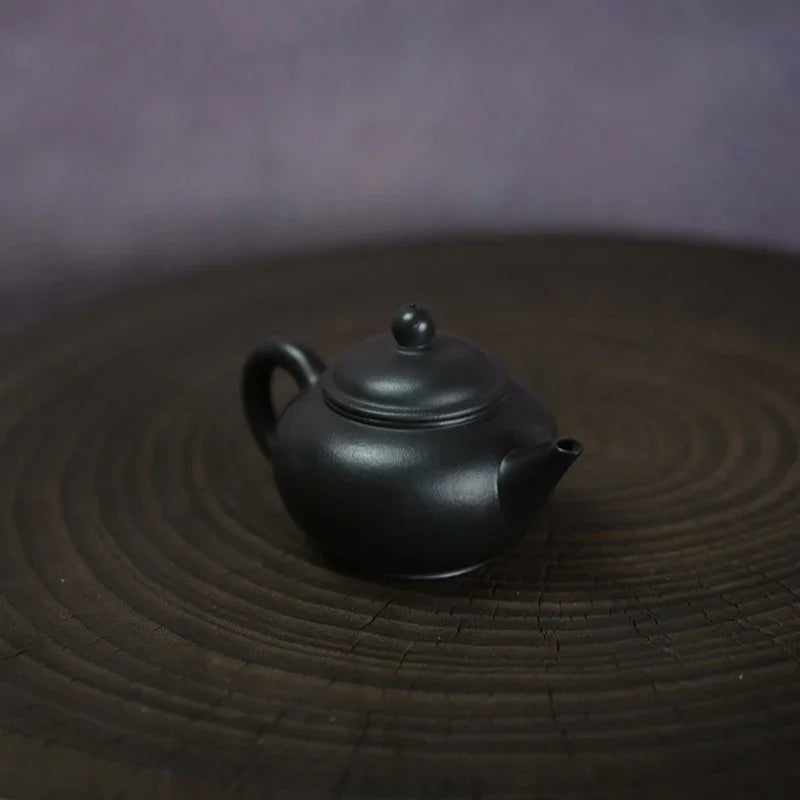 A small matte black ceramic teapot with a rounded body, loop handle, and short spout, placed on a dark circular wooden surface against a soft purple-gray background.

