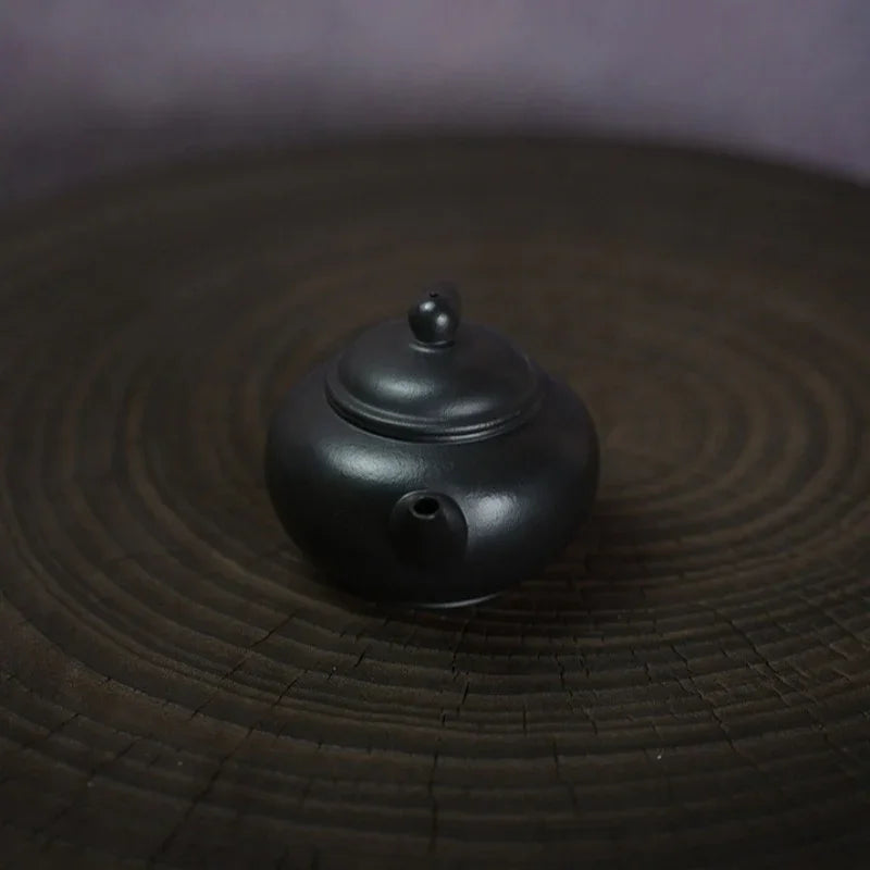 A small matte black ceramic teapot with a rounded body and lid, placed on a dark circular wooden surface.

