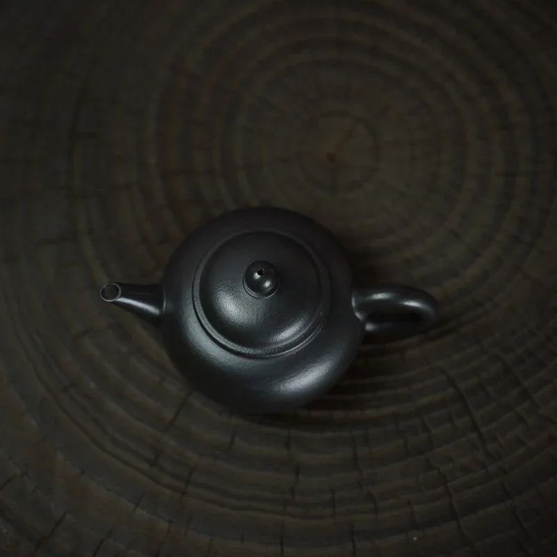 A matte black ceramic teapot with a rounded body, small spout, and loop handle, photographed from above on a dark wood grain surface.

