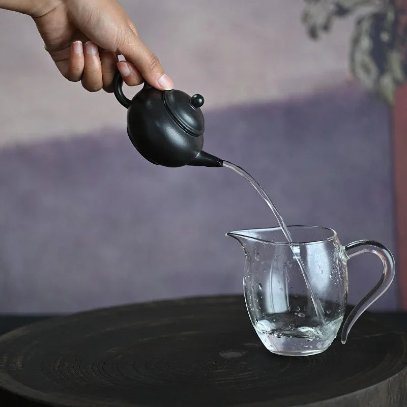 A hand holds a small matte black teapot pouring clear water into a transparent glass pitcher, set on a dark round tray against a soft blurred purple background.

