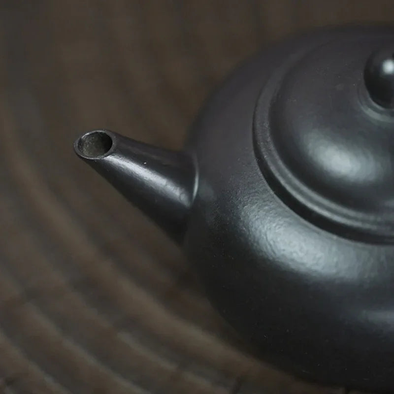 Close-up of a matte black ceramic teapot showing the spout and lid, resting on a dark wooden surface.

